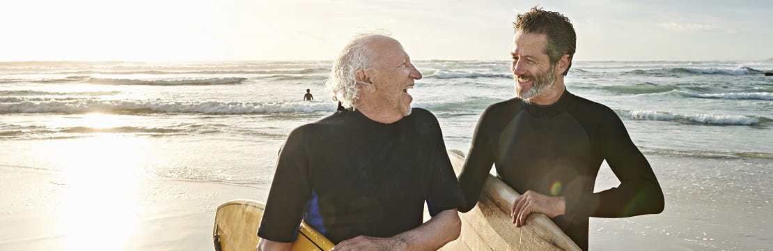 A father and son laugh together while carrying surfboards away from the beach.